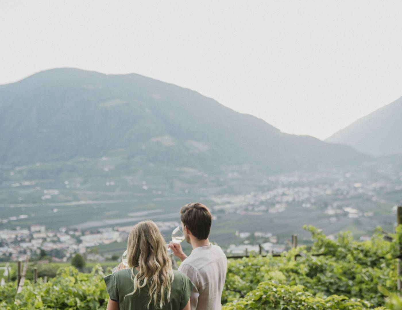 Pärchen genießt ein Glas Wein inmitten der Weinreben mit Blick auf Meran