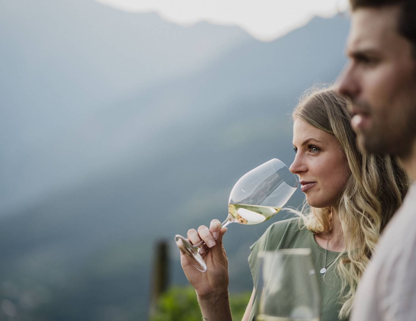 Pärchen genießt ein Glas Wein inmitten der Weinreben mit Blick auf Meran