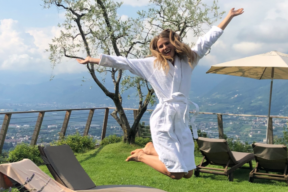 Fröhliche Frau im Bademantel springt auf der Terrasse des Wellnesshotels in Dorf Tirol
